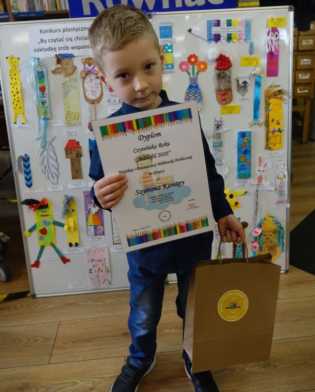 Chłopiec trzymający w ręku dyplom oraz torbę z nagrodami, w tle zakładki do książek