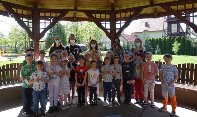 20 dzieci stoi pod zadaszeniem altany, za nimi dyrektorka biblioteki i bibliotekarka, 2 uczennice szkoły podstawowej oraz dyrektorka przedszkola