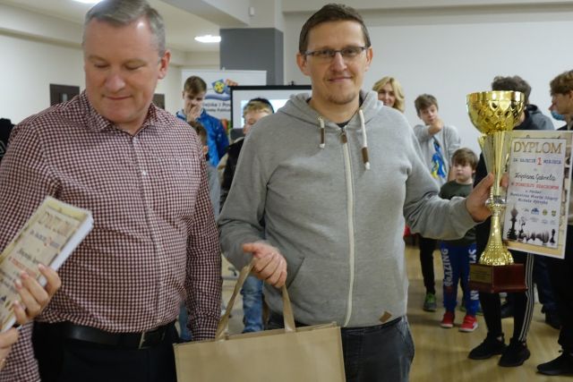 dwóch mężczyzn, po prawej w różowej koszuli w kratę, po prawej w siwej bluzie, trzymający nagrodę w torbie i w lewej dłoni puchar