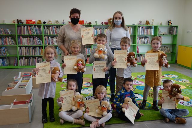 4 dzieci siedzi na zielonym dywanie, w rękach trzymają misia i dyplom, 5 dzieci stoi i trzyma misia i dyplom, za dziećmi stoją dwie osoby dorosłe