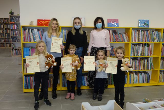 4 dzieci stoi, w rękach trzymają dyplom i misie, za nimi stoją 3 osoby dorosłe na tle żółtych regałów z książkami