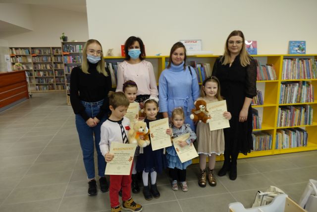 5 dzieci stoi, w rękach trzymają dyplom i misie, za nimi stoją 4 osoby dorosłe na tle żółtych regałów z książkami