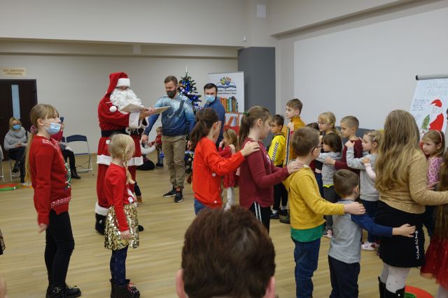 dzieci idą za sobą terzymając dłonie na ramionach dziecka przed sobą, w tyle osoby dorosłe, z lewej strony mikołaj