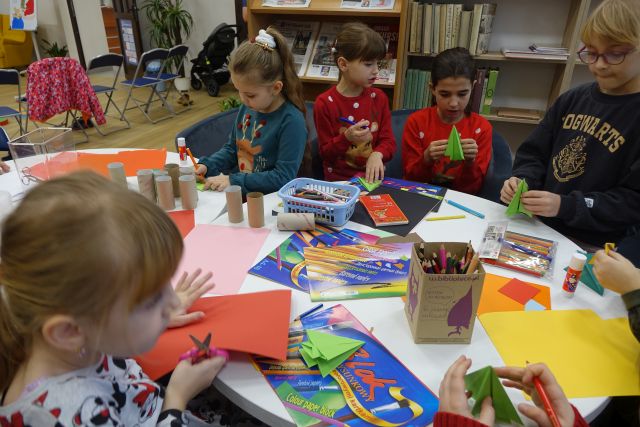 na okrągłym stole kolorowe kartki papieru, kredki, nożyczki, przy stole dzieci ozdabiające choinki 