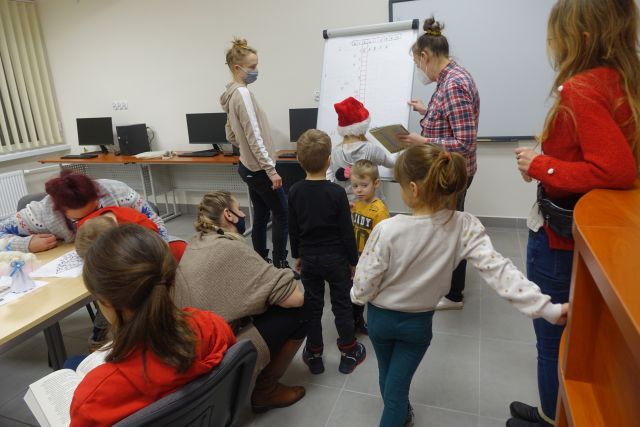 grupa dzieci i rodziców oraz bibliotekarka rozwiązują krzyżówkę na dużym białym papierze umieszczonym na flipcharcie