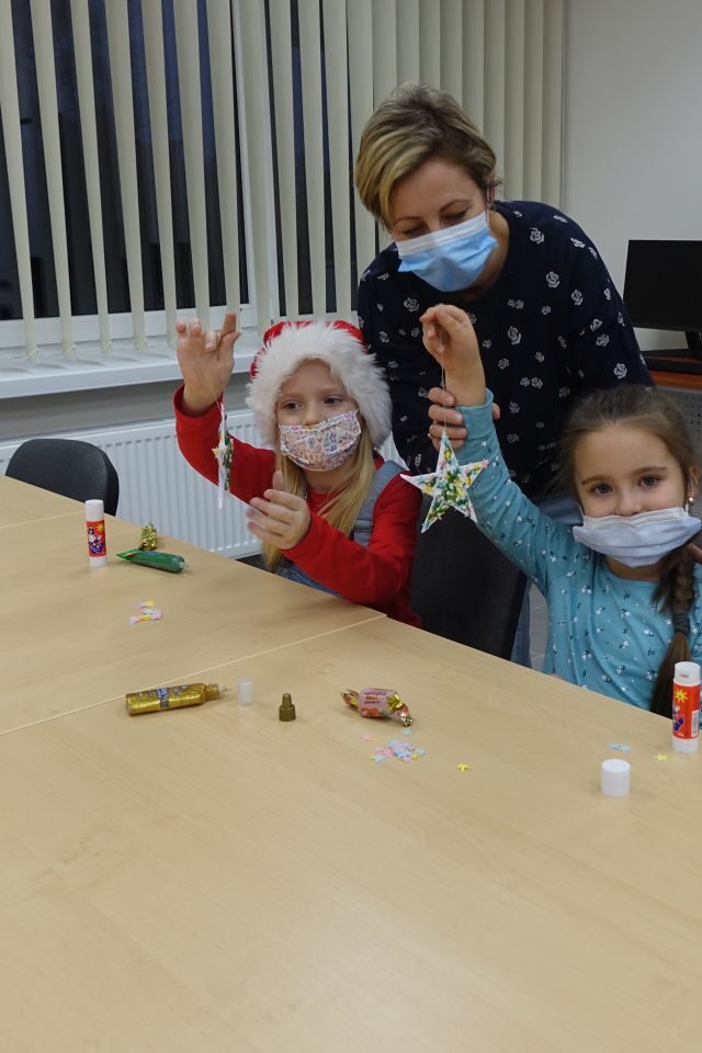 z przodu brązowy stól, za nim dwie dziewczynki trzymające w ręce gwiazdki, na ustach maseczki, za nimi pochylona kobieta w granatowej bluzce