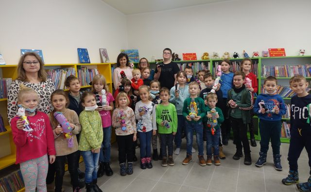 dzieci w kolorowych ubraniach stojące, za nimi 4 osoby dorosłe, za nimi żółte i zielone regały