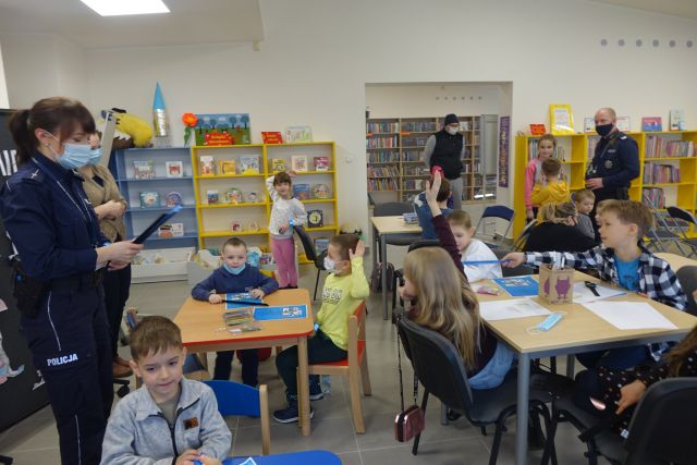 dzieci kolorowo ubrane siedzące przy brązowych stolikach, w koło przy ścianach kolorowe regały żółte, niebieskie, zielone, kobieta w stroju policyjnym niebieskim