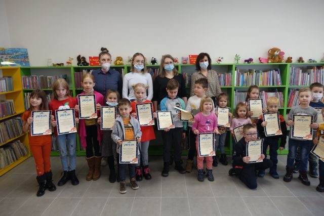 w dwóch rzędach dzici w kolorowych ubraniach stoją i trzymają w dłoniach dyplomy na jasnym papierze, za nimi stoją dorosłe 4 osoby kolorowo ubrane w maseczkach, za nimi żółte i zielone regały