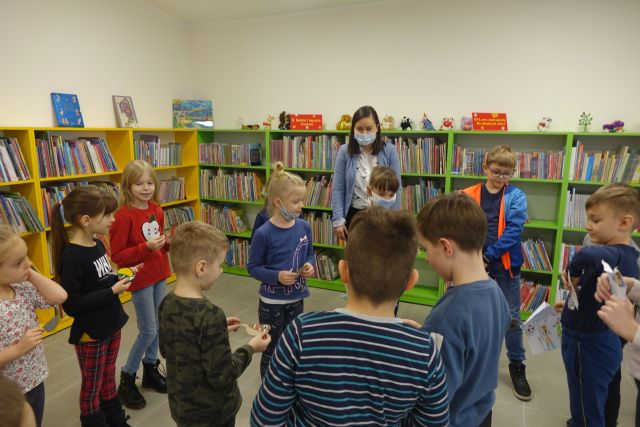 dzieci stojące w okręgu kolorowo ubrane, za nimi żółte i zielone regały