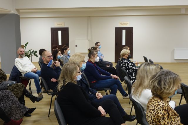 uczestnicy spotkania siedzący na krzesłach poubierani w ciemne stroje, z maseczkami na twarzach