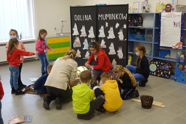 na podłodze kucają dzieci w kolorowych ubraniach, za nimi czarna tablica, a na niej białe muminki wycięte z papieru