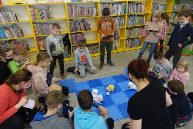 na podłodze rozłożona niebieska mata, na około siedzące i stojące dzieci w kolorowych ubraniach, na macie białe małe roboty, za dziećmi żółte regały