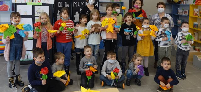 dzieci stoją i siedzą na podłodze w dwóch rzędach, kolorowo ubrani, trzymają w dłoniach kolorowe kwiaty z papieru