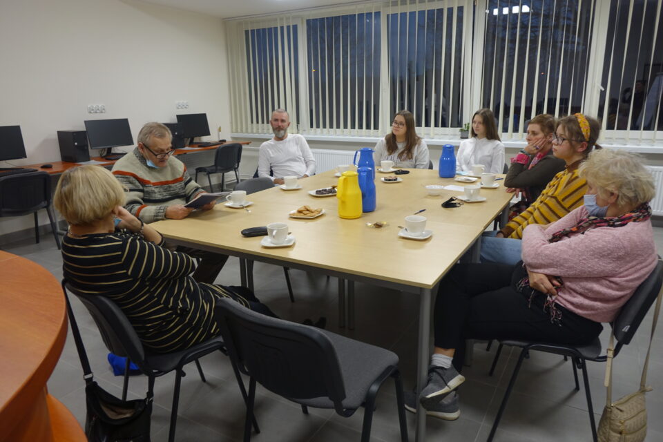 przy brązowych stołach wkoło siedzą na czarnych krzesłach kobiety i mężczyźni kolorowo ubrani, z tyłu okna z białymi roletami