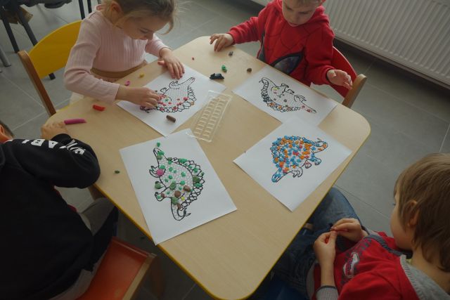 na brązowym stole białe kartki papieru, a na nich wyklejone kolorowe kulki z biblioły tworzące dinozaury, w koło stołu siedzi czwórka dzieci ubranych w kolorowe stroje