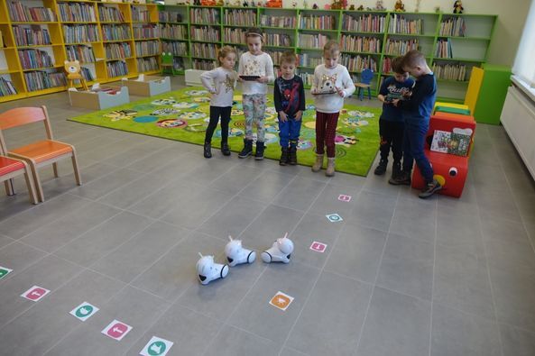 na siwej podłodze trzy małe białe roboty, za nimi stoi 6 dzieci w jasnych strojach, w dłoniach trzymają tablety, za nimi zielony dywan i zielone i żółte regały