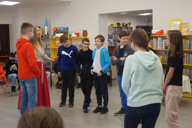 Młodzież w ciemnych i jasnych strojach stoi na środku sali, za nimi żółte regały