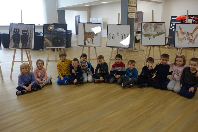 w tle sztalugi z obrazami przedstawiającymi zwierzęta: czarne koty, brązowe tygrysy i liski, biało-czarne pingwiny, czarne sowy na białym tle, na brązowej podłodze siedzą dzieci w kolorowych ubraniach