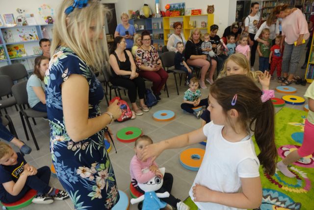na podłodze leży zielone dywan i okrągłe poduszki kolorowe do siedzenia, z tyłu na krzesłąch siedzą kobiety i mężczyźni, na środku stoją dzieci i dorosła kobieta w jasnych włosach