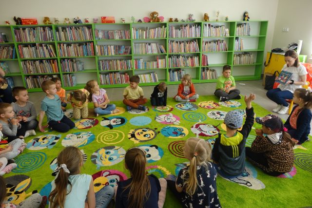 na zielonym dywanie siedzą dzieci w okręgu, za nimi zielone regały z książkami