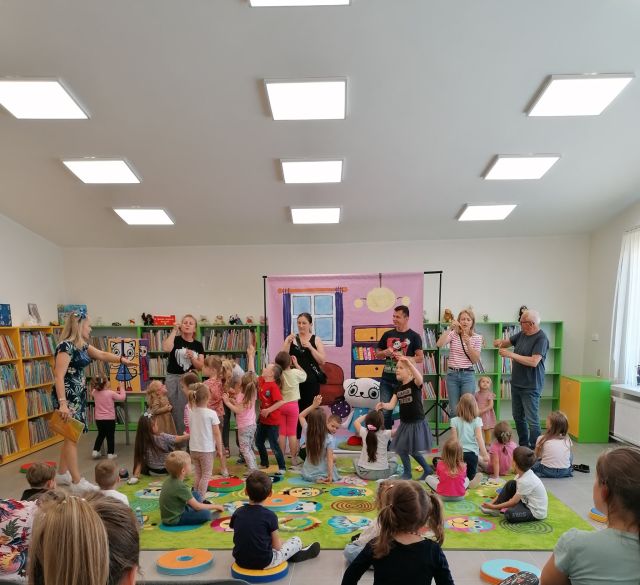 dzieci stoją i trzymają w górze dłonie, przed nimi kobieta, za nią plakat przedstawiający różowy pokój, z tyłu zielone regały