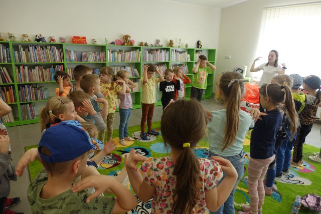 na zielonym dywanie stoją dzieci 2w okręgu, z prawej strony stoi kobieta w białej bluzce, za nią okna, za dziećmi zielone regały