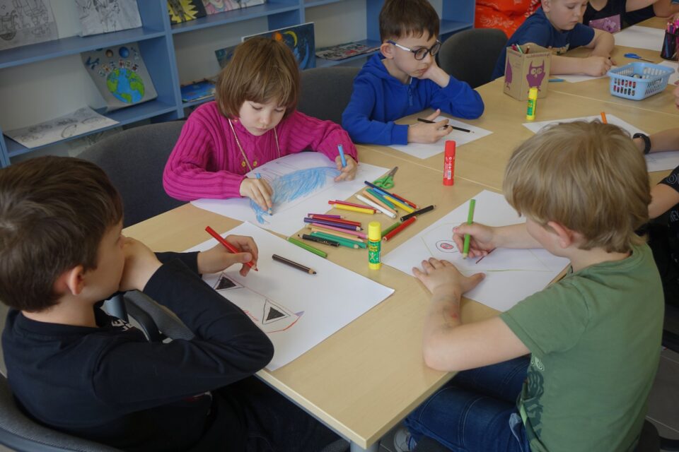 przy brązowych stołach siedzą kolorowo ubrane dzieci i malują na kartkach
