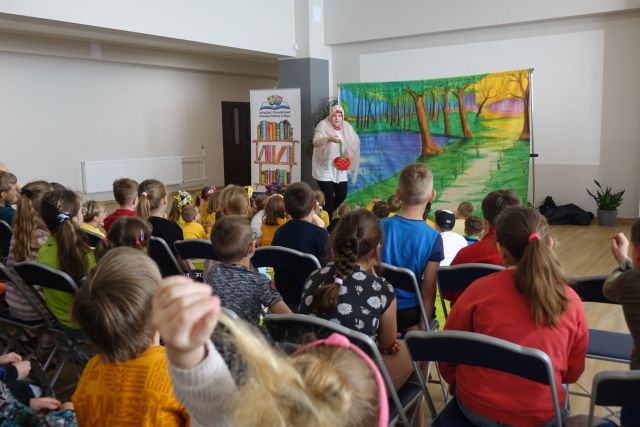 na niebieskich krzesłąch siedzą dzieci tyłem, przed nimi kobieta przebrana za czerwonego kapturka, w ręku trzyma czerwony koszyczek, za nią plakat lasu, z boku tablica z narysowanymi książkami