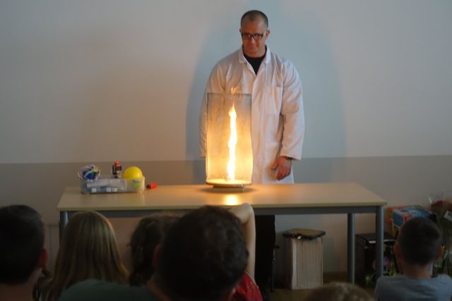 za brązowym stołem stoi mężczyzna w białym fartuchu, na stole stoi słój, a w nim pali się ogień