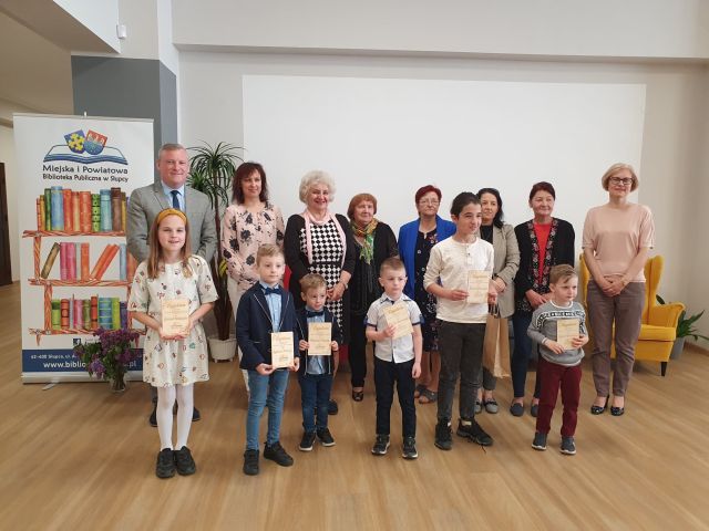 na tle białej ściany stoją w dwóch rzędach kobiety, mężczyźni i dzieci, wekach trzymają dyplomy