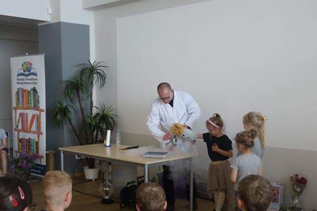 za brązowym stołęm stoi mężczyzna w białym faryuchu, z jego prawej strony dziewczynki, które pomagają w doświadczeniach, na rękach mnają duże rękawice