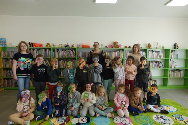 na zielonym dywanie stoją i siedzą dzieci, za nimi zielone regały z książkami