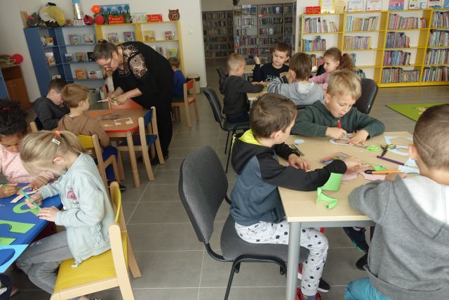 przy brązowych stolikach na siwych krzesłach siedzą dzieci, na stołaj mają kartki i wycinają z nich lupy i kolorują