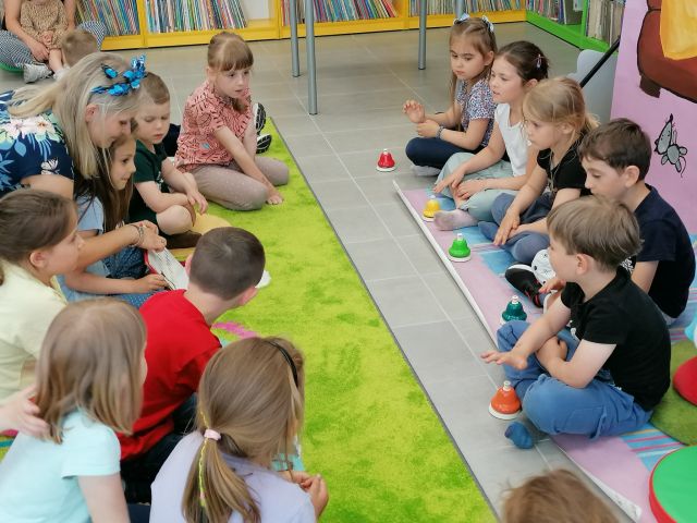 dzieci siedzą na zielonym dywanie, prze nimi nsiedzi 5 dzieci, przed nimi instrumenty do grania