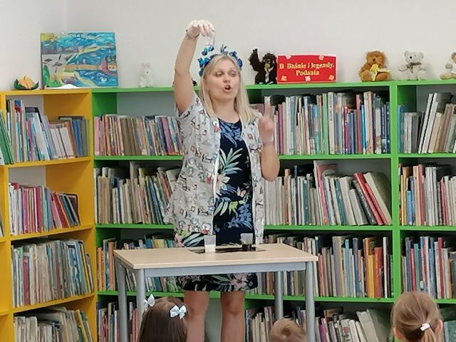 przy stoliku stoi kobieta w fartuchu białym w kwiatki, w uniesionej dłoni trzyma kubek z płynem, za nią żółte i zielone regały z książkami