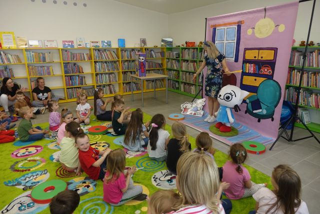 na zielonym dywanie siedzą dzieci, przed nimi stoi kobieta w niebieskiej sukience, z boku żółte regały, z tyłu zielone regały