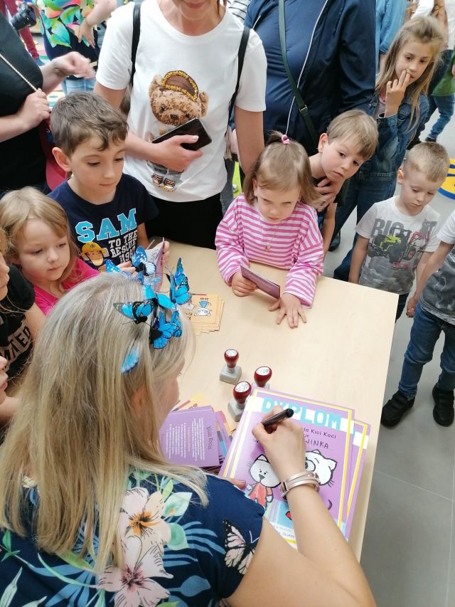 przy brązowym stoliku siedzi kobieta i wypisuje niebieski dyplom z kotem, wokół stolika stoją dzieci