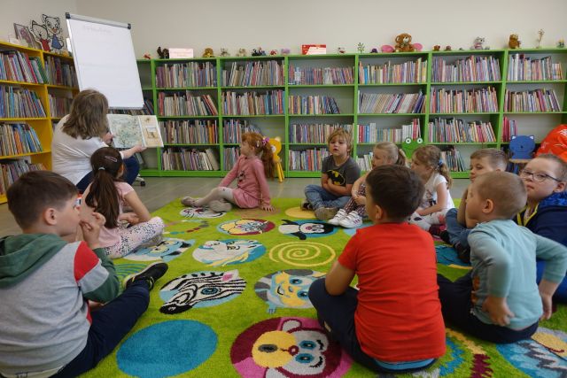 na zielonym dywanie siedzą dzieci, przed nimi na krzesełku siedzi kobieta i trzyma w dłoniach książkę, za nimi zielone regały z książkami, a po lewej stronie żółte regały