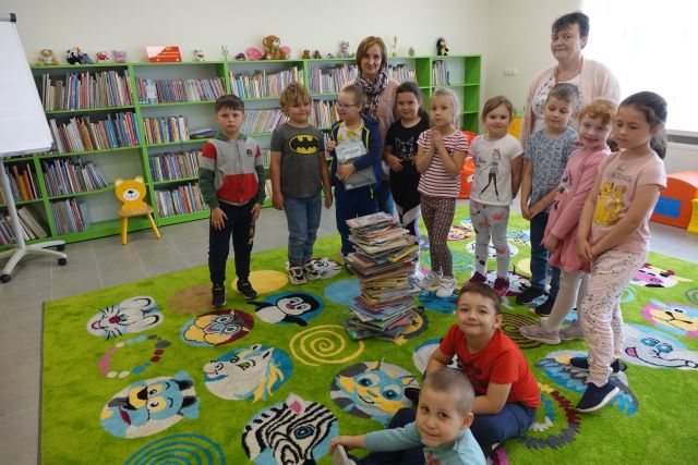 na zielonym dywanie stoją i siedzą dzieci oraz 2 osoby dorosłe, na środku dywanu góra z książek, za nimi zielone regały z książkami