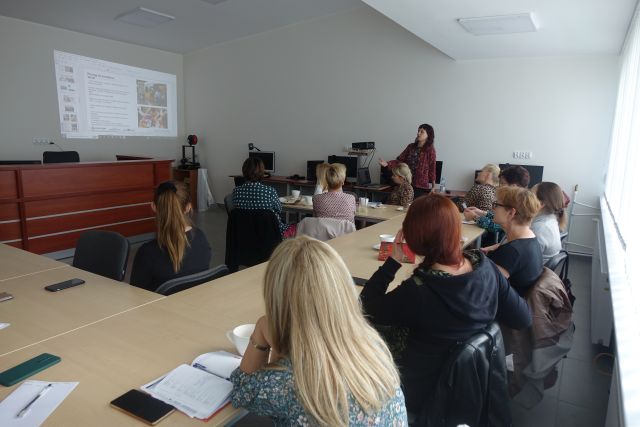 uczestnicy szkolenia siedzą przy stołąch, przed nimi stoi kobieta w bordowej bluzce w wzory