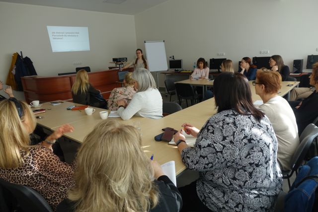 uczestnicy szkolenia siedzą przy stołach, przed nimi stoi prowadząca, w tle ekran z prezentacją
