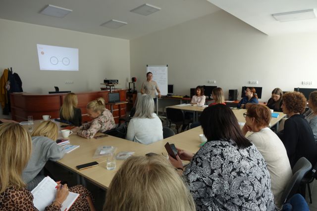 uczestnicy szkolenia siedzą przy stołach, przed nimi stoi prowadząca, w tle ekran z prezentacją