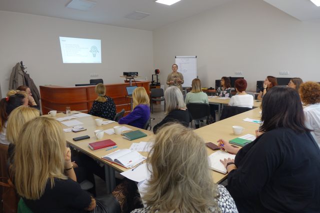 uczestnicy szkolenia siedzą przy stołach, przed nimi stoi prowadząca, w tle ekran z prezentacją