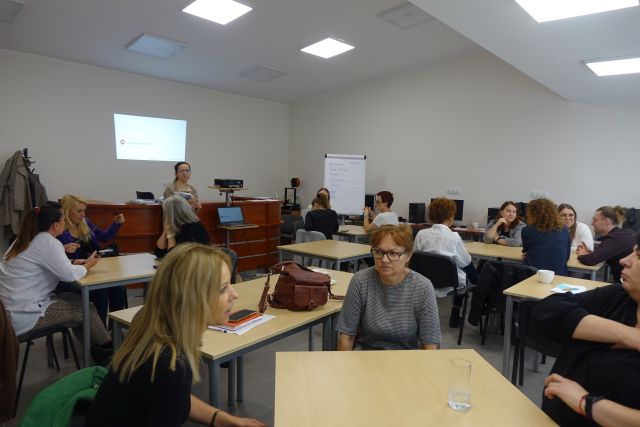 uczestnicy szkolenia siedzą przy stołach, przed nimi stoi prowadząca, w tle ekran z prezentacją