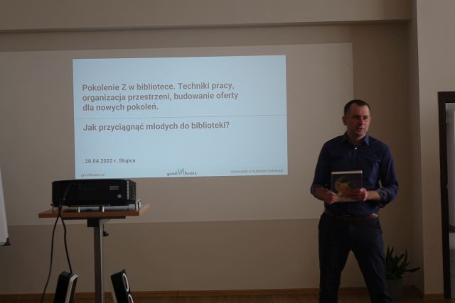 Na na środku jasnej ściany biały ekran, przed nim brązowy stolik, na nim rzutnik czarny, z prawej strony stoi mężczyzna w granatowej koszuli i czarnych spodniach, w rękach trzyma książkę