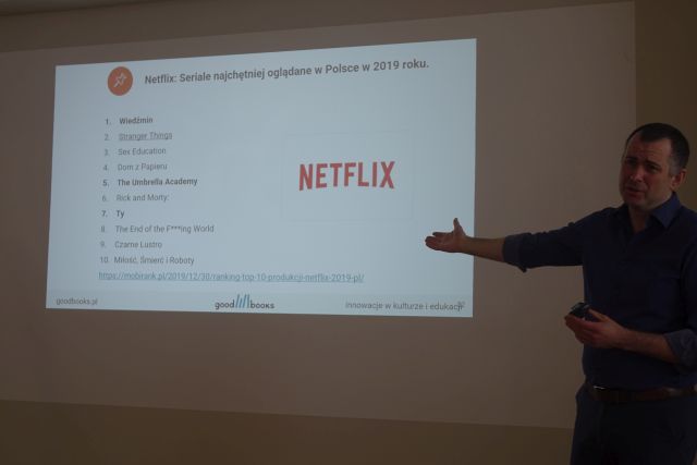 na szarej ścianie biały ekran, z prawej strony mężczyzna w granatowej koszuli i czarnych spodniach z wyciągniętą w bok ręką z lewej strony