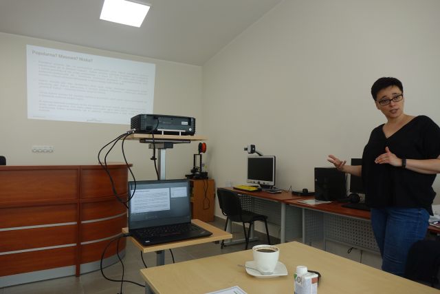 kobieta w czarnej bluzce i jasnych dżinsach stoi przed uczesnikami szkolenia, w tle biurko, stolik z laptopem i rzutnik