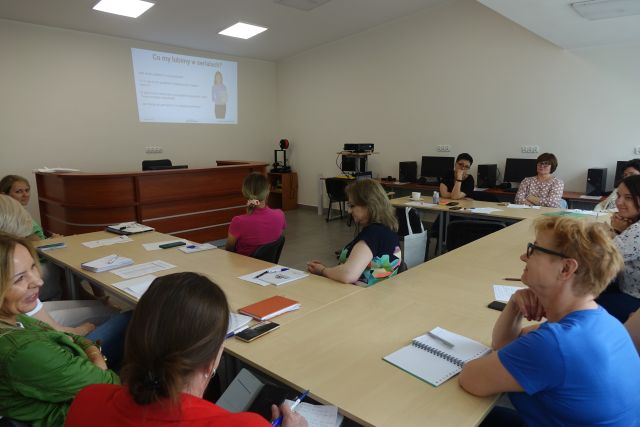 uczesniczki szkolenia siedzą przy stołach, na stołąch mają zeszyty, w tle brązowe biurko, rzutnik na stoliku