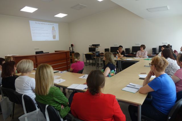 uczestnicy szkolenia siedzą przy stołąch, w tle biurko, rzutnik na stoliku, na ścianie ekran z prezentacją
